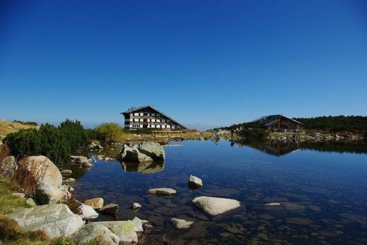 Bezbog Hut reflected in the clear waters of Bezbog Lake, with rocks along the shore in the Pirin Mountains