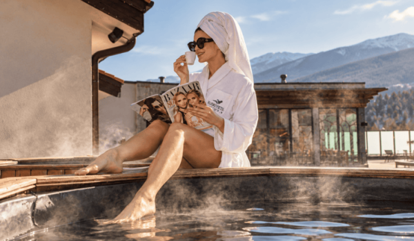 Woman in a bathrobe enjoying coffee beside a steaming outdoor hot tub at Ruskovets Resort, with snow-capped mountains behind her
