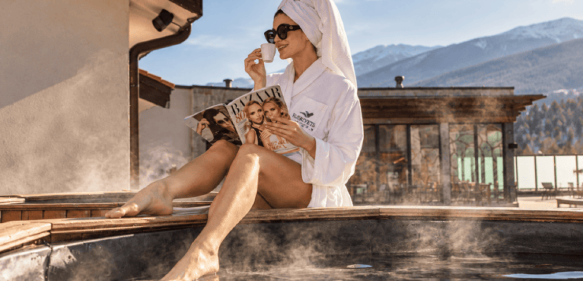 Woman in a bathrobe enjoying coffee beside a steaming outdoor hot tub at Ruskovets Resort, with snow-capped mountains behind her